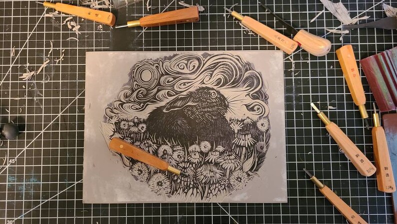 May include: A black and white linocut print of a rabbit in a field of dandelions. The rabbit is looking to the right, and the moon is visible in the sky above. The print is surrounded by carving tools on a black grid.