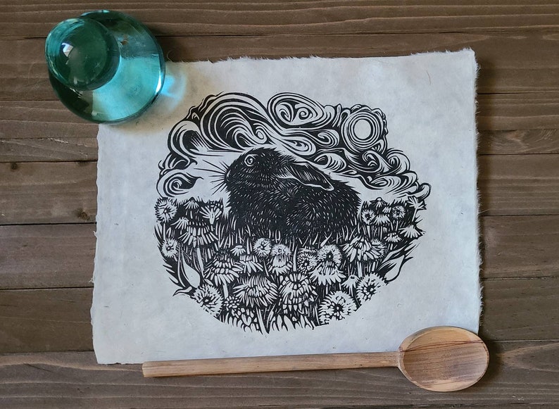 May include: Black and white linocut print of a rabbit in a field of flowers. The rabbit is looking to the right and the flowers are in bloom. The print is on a white background.