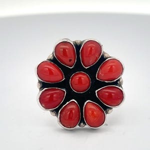 May include: A silver ring featuring a flower design with eight vibrant red teardrop-shaped stones surrounding a central red stone. The ring has a dark background and a polished silver band.