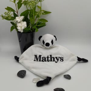 Peut inclure: Une couverture de sécurité en peluche blanche avec une tête de panda aux oreilles et aux yeux noirs. Le centre du tissu doux est brodé du nom Mathys en fil noir. La couverture présente de petits membres en peluche noire sur les côtés.