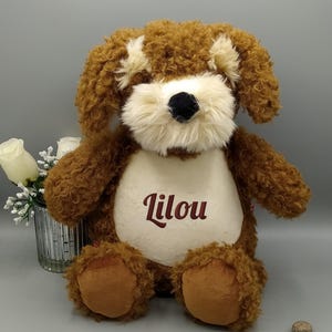 Peut inclure: Une peluche en forme de chien marron avec des poils bouclés, un museau crème et un nez noir. Le jouet porte le nom "Lilou" brodé sur sa poitrine. Le chien est posé sur une surface grise.