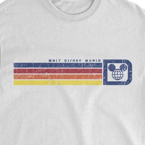 May include: White t-shirt featuring a retro design with horizontal stripes in blue, red, orange, and yellow. The text "WALT DISNEY WORLD" is above the stripes, with a Mickey Mouse head logo on the right. A vintage-style graphic tee.