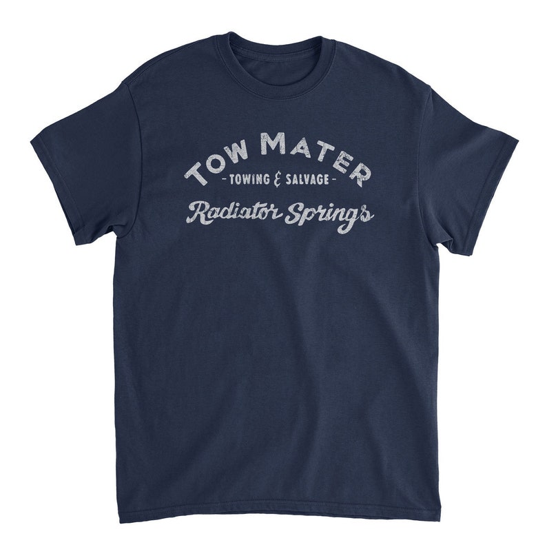 Pu&ograve; includere: Una t-shirt blu navy con una grafica bianca in stile vintage che recita "Tow Mater - Towing & Salvage - Radiator Springs".