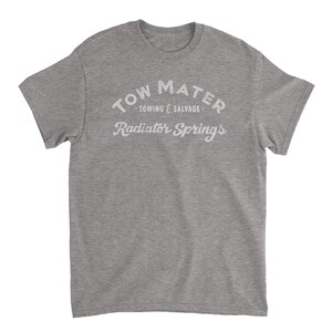 Pu&ograve; includere: Una t-shirt grigia melange con una stampa bianca che dice "Tow Mater - Towing & Salvage - Radiator Springs".