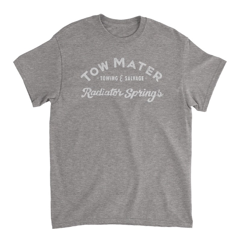 Pu&ograve; includere: Una t-shirt grigia melange con una stampa bianca che dice "Tow Mater - Towing & Salvage - Radiator Springs".
