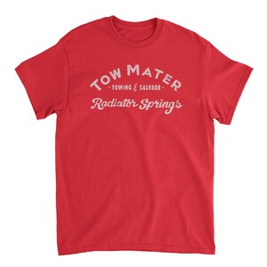Pu&ograve; includere: Maglietta rossa con testo bianco che recita "Tow Mater - Towing & Salvage - Radiator Springs".