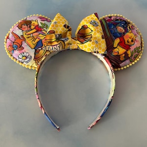 May include: A whimsical headband featuring Winnie the Pooh and friends. The ears are decorated with colorful illustrations of the characters, and the bow has a honeycomb pattern. The edges are trimmed with yellow pom-poms, adding a playful touch to this accessory.