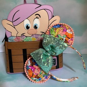 May include: A brown faux leather backpack with a cartoon image of a dwarf with pink hair peeking out of the top. The backpack has a wooden barrel design and a green sequin bow with a floral print on the side. The backpack is paired with a set of matching Minnie Mouse ears with a green sequin bow and a floral print.