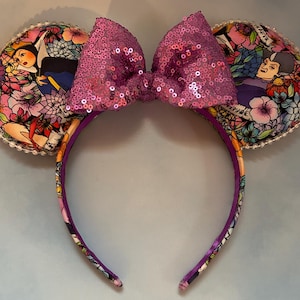 May include: A headband with two ear-shaped decorations and a large sequined bow. The ears feature a colorful floral pattern with images of characters. The headband and ears are trimmed with a white beaded edge.