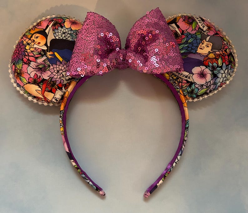 May include: A headband with two ear-shaped decorations and a large sequined bow. The ears feature a colorful floral pattern with images of characters. The headband and ears are trimmed with a white beaded edge.