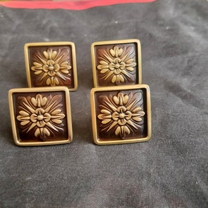 May include: Four square-shaped cabinet knobs with a gold finish and a brown center. Each knob features a raised, ornate floral design in a lighter gold color.