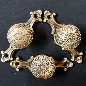 May include: Three ornate brass cabinet knobs with a starburst design. Each knob has a decorative scrollwork base.