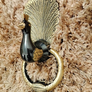 Könnte beinhalten: Ein dekorativer Türklopfer mit Pfauendesign. Der Klopfer hat einen schwarzen Körper mit goldenen Akzenten, einschließlich eines detaillierten Schwanzes und eines ringförmigen Griffs. Der Hintergrund ist eine strukturierte, braune Oberfläche.