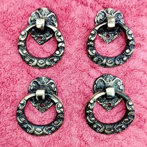 Könnte beinhalten: Vier antike bronzefarbene Schrankgriffe mit kunstvollen, wirbelnden Designs. Jeder Griff hat eine dekorative Rückplatte und einen kreisförmigen Griff. Die Hardware befindet sich auf einem rosa, strukturierten Hintergrund.