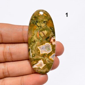 May include: Oval shaped green, brown, white, and orange moss agate cabochon. The stone has a polished finish and is approximately 5 cm long.