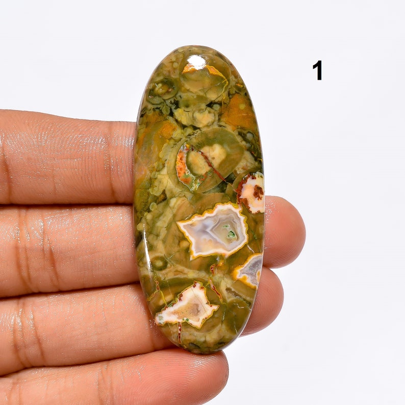 May include: Oval shaped green, brown, white, and orange moss agate cabochon. The stone has a polished finish and is approximately 5 cm long.