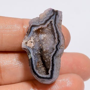 May include: A natural geode specimen with a cavity lined with sparkling crystals. The exterior features bands of gray, black, and white, creating a layered effect. The geode is held in a hand, showcasing its size and texture.