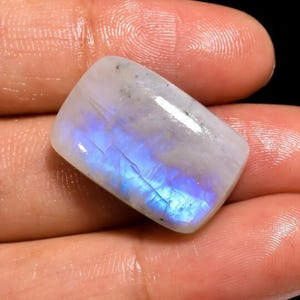Könnte beinhalten: Ein weißer Mondstein-Cabochon mit einem blauen, irisierenden Schimmer. Der Stein ist rechteckig und hat eine glatte, polierte Oberfläche.