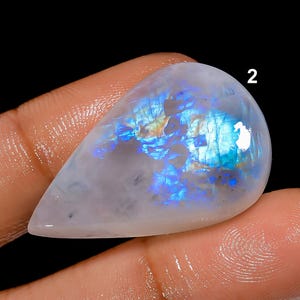 May include: A pear-shaped, polished moonstone gemstone. The stone has a translucent, milky white base with iridescent blue and gold flashes. The number "2" is visible on the top right of the stone. The gemstone is held between two fingers against a black background.