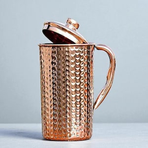 Puede incluir: Una jarra de color cobre con acabado martillado y tapa. La jarra tiene forma cilíndrica con un asa curvada. La tapa está ligeramente entreabierta, revelando el interior. La textura martillada le da a la jarra un aspecto rústico pero elegante.