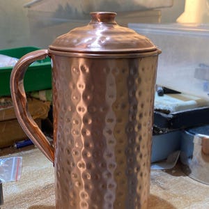 May include: A copper-colored pitcher with a lid and a handle. The pitcher has a hammered texture and a rounded shape. The handle is curved and attached to the side of the pitcher. The lid has a small knob on top.