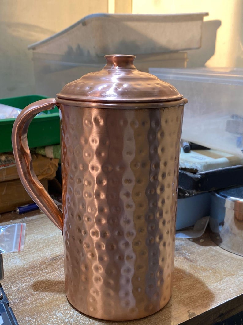 May include: A copper-colored pitcher with a lid and a handle. The pitcher has a hammered texture and a rounded shape. The handle is curved and attached to the side of the pitcher. The lid has a small knob on top.