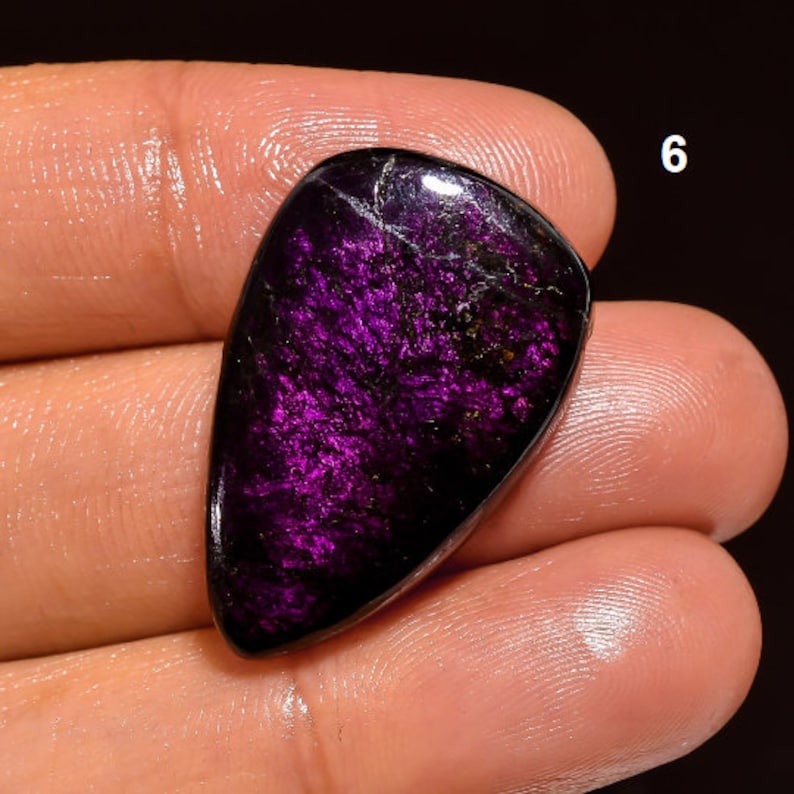 May include: A dark purple and black gemstone cabochon with a teardrop shape. The stone has a shimmery, iridescent finish.