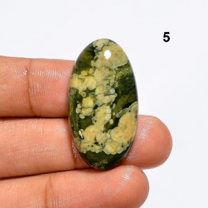 May include: Oval shaped green and white moss agate cabochon with a swirling pattern. The stone is approximately 5 cm long.