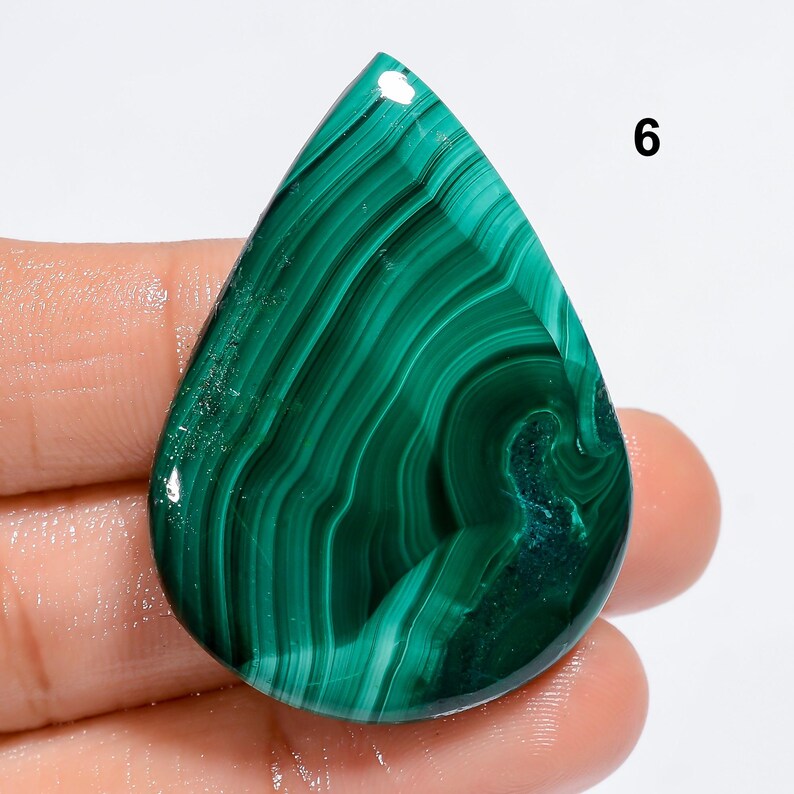 May include: A green malachite cabochon gemstone with a swirling pattern. The stone is cut in a teardrop shape and has a smooth, polished surface.