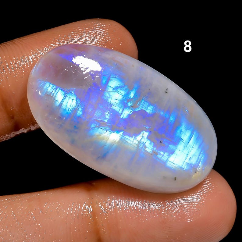 May include: An oval-shaped, polished moonstone gemstone. The stone has a milky white base with iridescent blue and purple flashes. The number "8" is visible in white text in the upper right corner. The gemstone is held between two fingers.