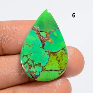 May include: A green and brown turquoise cabochon gemstone in a teardrop shape. The stone has a mottled pattern and is set in a silver bezel.