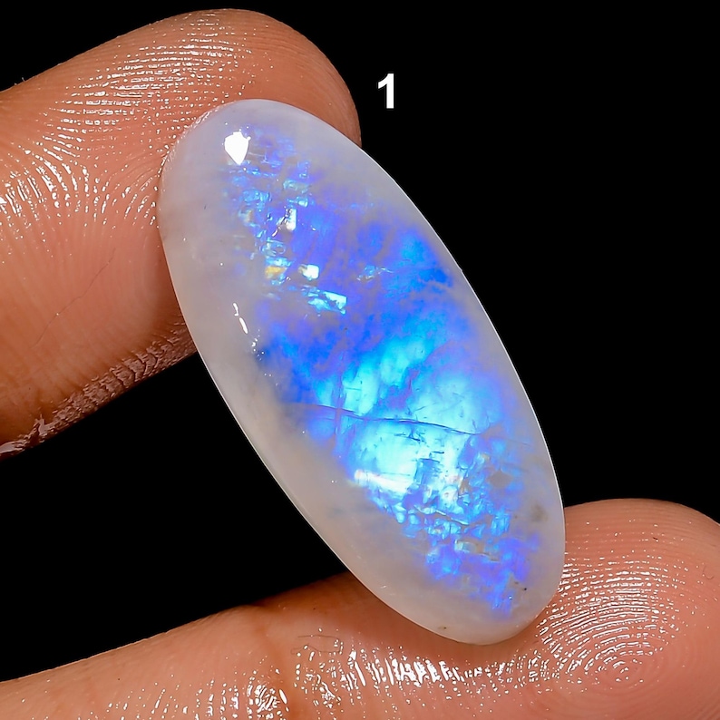May include: A polished, oval-shaped gemstone with a milky white base and iridescent blue and gold flashes. The gemstone is held between fingers against a black background. The number "1" is visible at the top of the image.