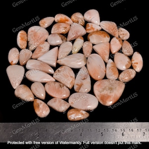 Op de afbeelding: Een verzameling van 25 roze en oranje edelsteen cabochons, in verschillende vormen en maten. De cabochons zijn gerangschikt op een zwarte ondergrond met een liniaal voor schaal.