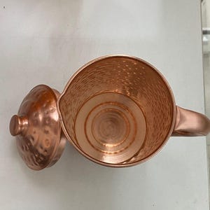 May include: A copper-colored pitcher with a matching lid. The pitcher has a hammered texture and a handle. The lid is off to the side, revealing the interior of the pitcher. The pitcher is likely used for serving beverages.