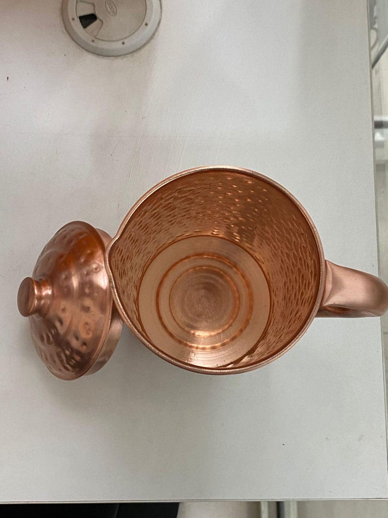 May include: A copper-colored pitcher with a matching lid. The pitcher has a hammered texture and a handle. The lid is off to the side, revealing the interior of the pitcher. The pitcher is likely used for serving beverages.