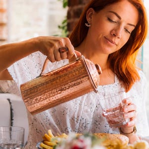 Puede incluir: Una jarra de cobre martillado con cuerpo cilíndrico y tapa a juego, utilizada para verter agua en un vaso transparente. La jarra presenta un acabado metálico con una superficie texturizada y un asa lateral resistente y firme.