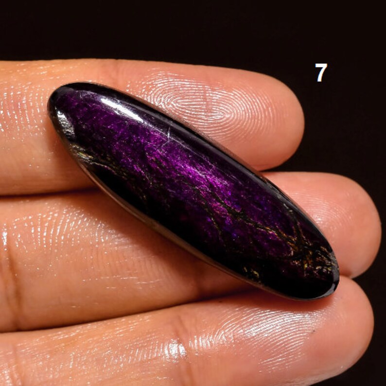 May include: A purple and black oval-shaped gemstone with a shimmering, iridescent finish. The stone is held in a hand, and the number "7" is visible in the background.