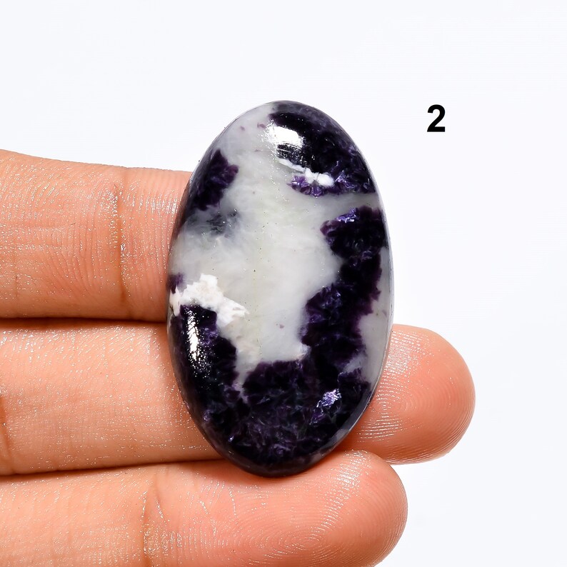 May include: Oval shaped cabochon gemstone with a white and purple pattern. The stone is polished and has a glossy finish.