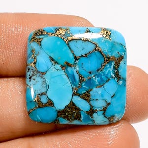 May include: A square-shaped turquoise gemstone with a polished surface. The stone features a vibrant blue color with intricate patterns of brown and gold veining throughout. The gemstone is held in a hand.