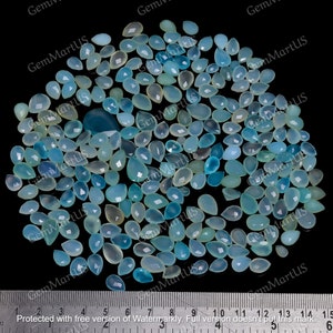 Può includere: Un grande gruppo di pietre preziose a forma di lacrima di colore blu chiaro e bianco, alcune con un colore bianco più opaco, sono sparse su una superficie nera. Un righello è visibile nella parte inferiore dell'immagine, con segni da 1 a 15.