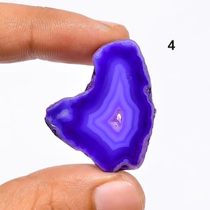 Puede incluir: Una rebanada de ágata púrpura con bandas blancas. La ágata es translúcida y tiene una forma natural irregular.