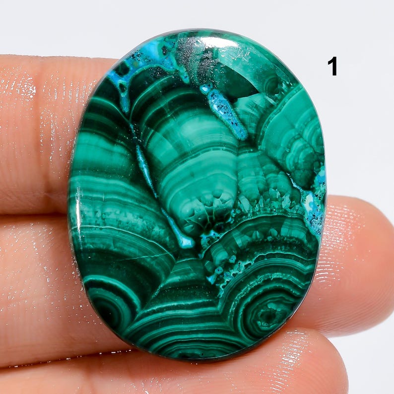May include: Oval shaped green and blue malachite cabochon gemstone with a swirling pattern. The stone is approximately 2 inches long and 1.5 inches wide.