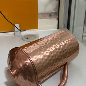 May include: A copper-colored pitcher with a hammered texture and a matching lid. The pitcher has a curved handle and a rounded knob on the lid. The overall design is simple and elegant, suitable for serving beverages.