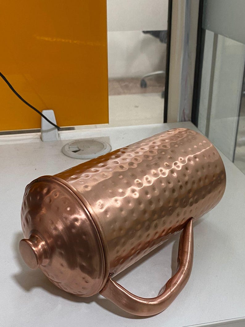 May include: A copper-colored pitcher with a hammered texture and a matching lid. The pitcher has a curved handle and a rounded knob on the lid. The overall design is simple and elegant, suitable for serving beverages.