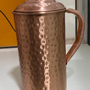 May include: A copper-colored pitcher with a lid and handle. The pitcher has a hammered texture and a rounded base. The handle is curved and attached to the side of the pitcher. The lid sits on top of the pitcher and has a small knob.