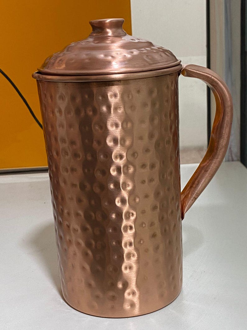May include: A copper-colored pitcher with a lid and handle. The pitcher has a hammered texture and a rounded base. The handle is curved and attached to the side of the pitcher. The lid sits on top of the pitcher and has a small knob.