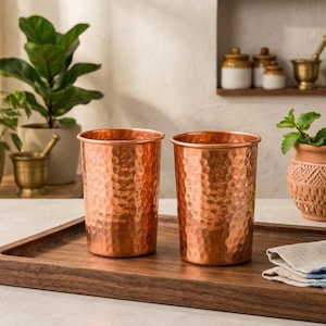 Puede incluir: Dos vasos cilíndricos fabricados en cobre pulido, con una textura martillada distintiva en toda la superficie exterior. Las tazas tienen un borde liso y plano, con un acabado metálico de color naranja parduzco que refleja la luz.