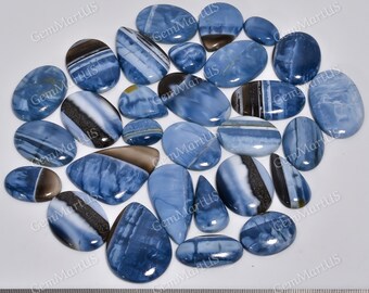 May include: A collection of 20 blue opal cabochons, each with unique patterns and markings. The cabochons are oval and teardrop shaped, and range in size from approximately 1 to 2 inches in length. The cabochons are arranged on a white surface with a ruler for scale.
