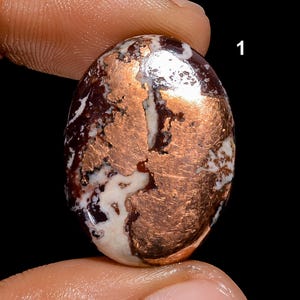 Może przedstawiać: Owalny kamień szlachetny w szlifie kaboszonowym z wzorem miedzianym i białym. Kamień ma gładką, wypolerowaną powierzchnię.
