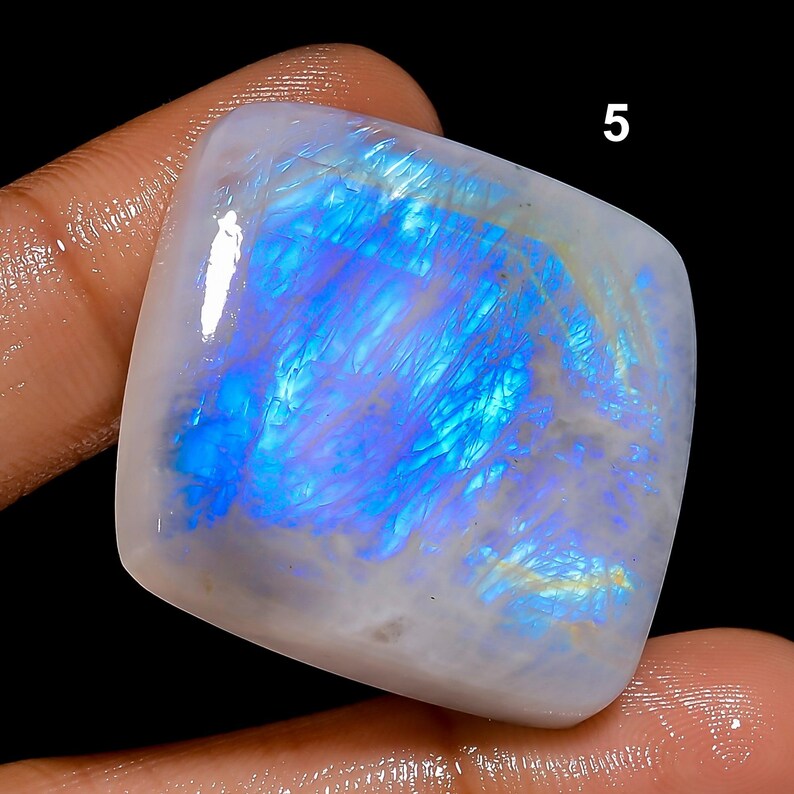May include: A square-cut, polished gemstone with a pearly white base and iridescent blue and yellow flashes. The number "5" is visible in the upper right corner. The stone is held by a person's fingers.